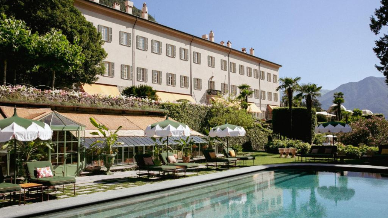 Un lussuoso hotel COMO con una facciata chiara e numerose finestre si affaccia su un lussureggiante giardino pieno di lettini, ombrelloni, palme e una piscina limpida sotto un cielo azzurro brillante con montagne in lontananza.