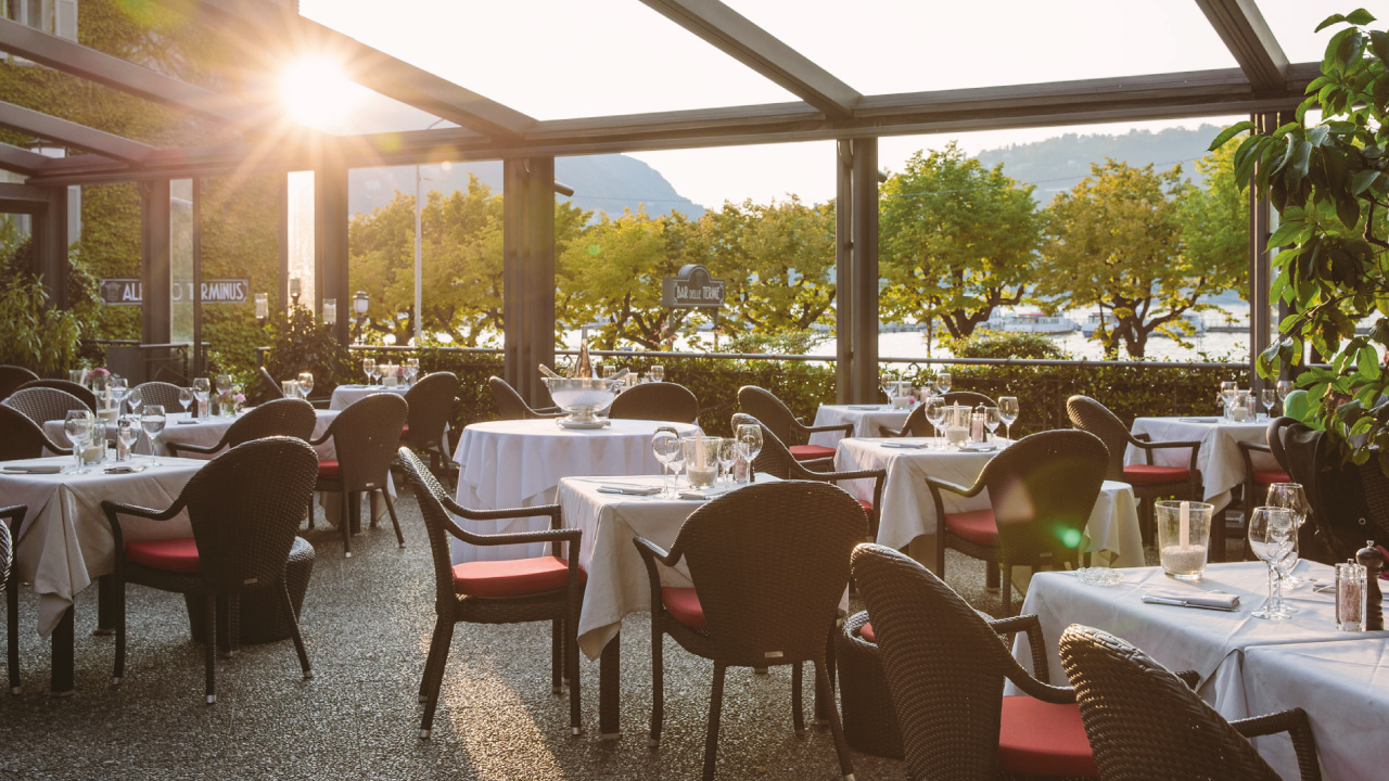 Un patio all'aperto con tavoli rotondi coperti da tovaglie bianche e sedie in tessuto intrecciato scuro, apparecchiato per la cena. La luce del sole filtra attraverso una tettoia in vetro, con alberi verdi e una vista sull'acqua che ricorda il Lago di Como sullo sfondo.