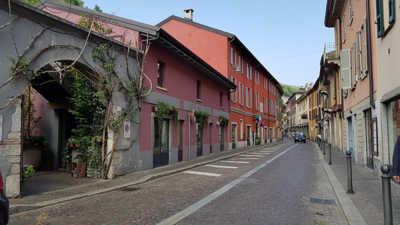 Una tranquilla strada acciottolata di una città europea, molto simile a quelle di Como, è fiancheggiata da edifici colorati, piante in vaso e una vegetazione lussureggiante. Un'auto è parcheggiata in lontananza; il cielo sopra di noi è limpido e azzurro.