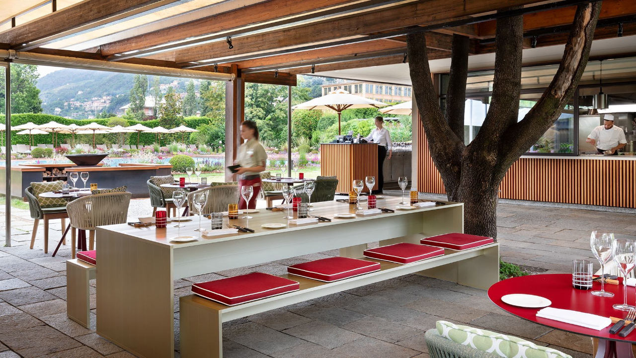 Un ristorante all'aperto con arredi moderni, un lungo tavolo con panche imbottite di rosso apparecchiato per la cena: il personale serve gli ospiti mentre gli chef lavorano in una cucina a vista. Alberi di Como e vegetazione circondano l'area, con le montagne visibili sullo sfondo.