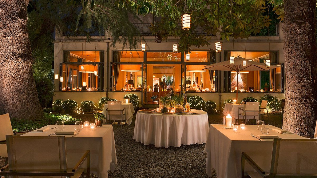 Patio del ristorante all'aperto di sera, con tavoli rotondi elegantemente apparecchiati, tovaglie bianche, candele e lucine, circondato da alberi: proprio come in una romantica serata a Como. Una luce calda filtra dalle ampie finestre dell'edificio sullo sfondo.