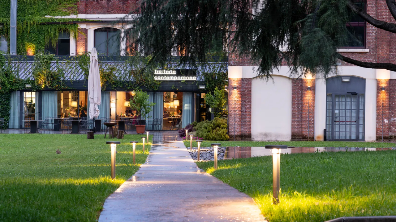 Un sentiero illuminato attraversa un prato verde e conduce all'ingresso di un ristorante con ampie finestre, posti a sedere all'aperto e pareti in mattoni, circondato dal verde e da luci soffuse la sera, proprio come in una scena di Como.