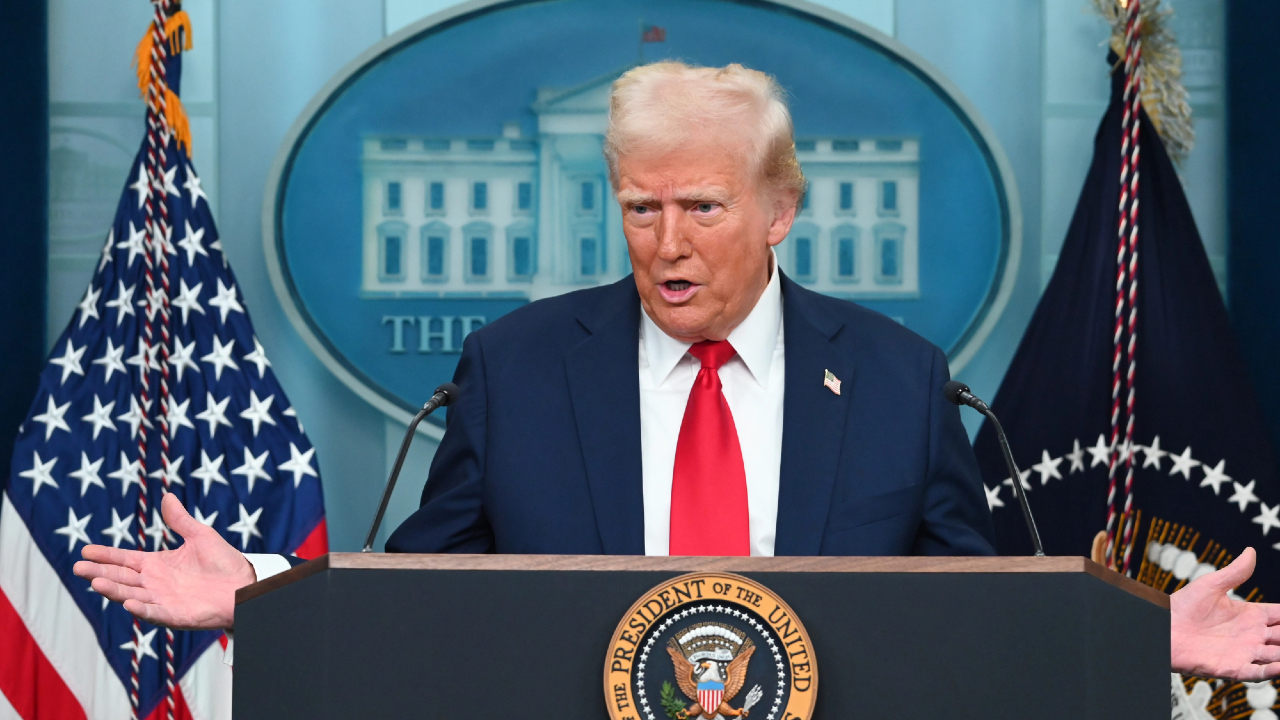 Un uomo in abito blu navy e cravatta rossa è in piedi su un podio con il sigillo presidenziale degli Stati Uniti, gesticolando mentre discute di azioni chiave. Sullo sfondo sono visibili le bandiere americane e l'emblema della Casa Bianca.