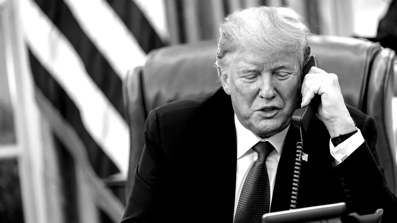 Un uomo in giacca e cravatta è seduto a una scrivania e parla al telefono fisso. Sullo sfondo si vede una bandiera americana. L'immagine è in bianco e nero.