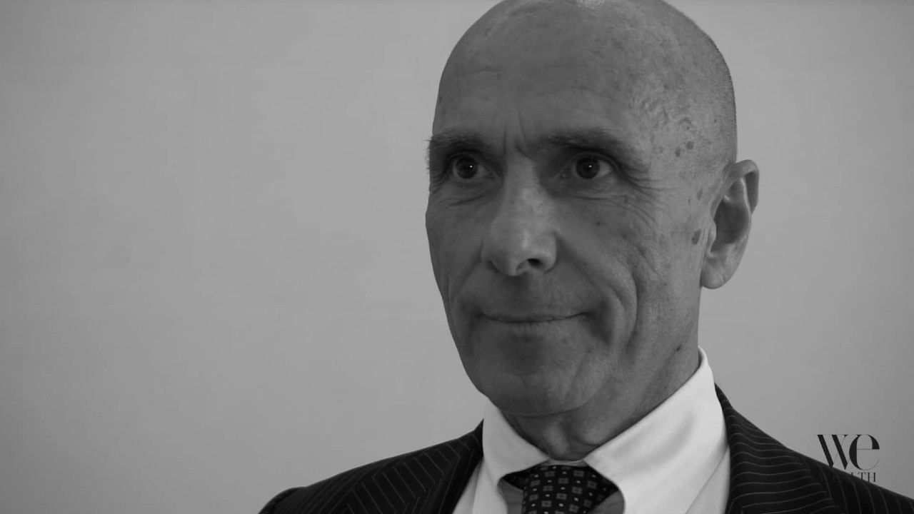 Foto in bianco e nero di un uomo anziano in giacca e cravatta, con sguardo leggermente laterale e un'espressione neutra. Lo sfondo semplice e il piccolo logo "WE" nell'angolo in basso a destra conferiscono un classico stile da ritratto TNB.