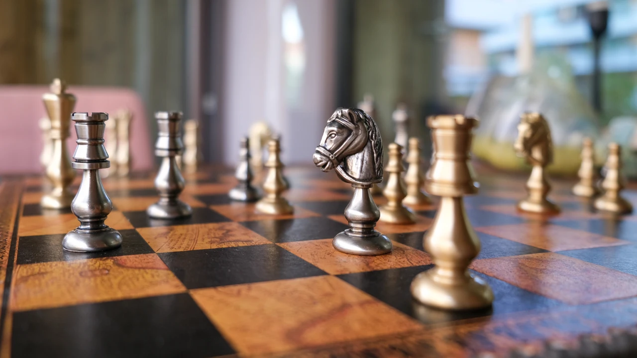 Primo piano di una scacchiera con pezzi d'argento e oro e un cavallo al centro. La luce naturale e soffusa ne esalta i dettagli, mentre gli altri pezzi sono sfocati. Il riferimento va alle strategie usate da imprese e Hnwi su come investire tra dazi e guerre fiscali.