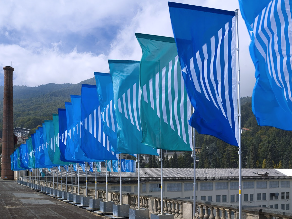 File di bandiere blu e verde acqua con strisce bianche diagonali sventolano nel vento lungo un balcone, con montagne ed edifici industriali sullo sfondo.