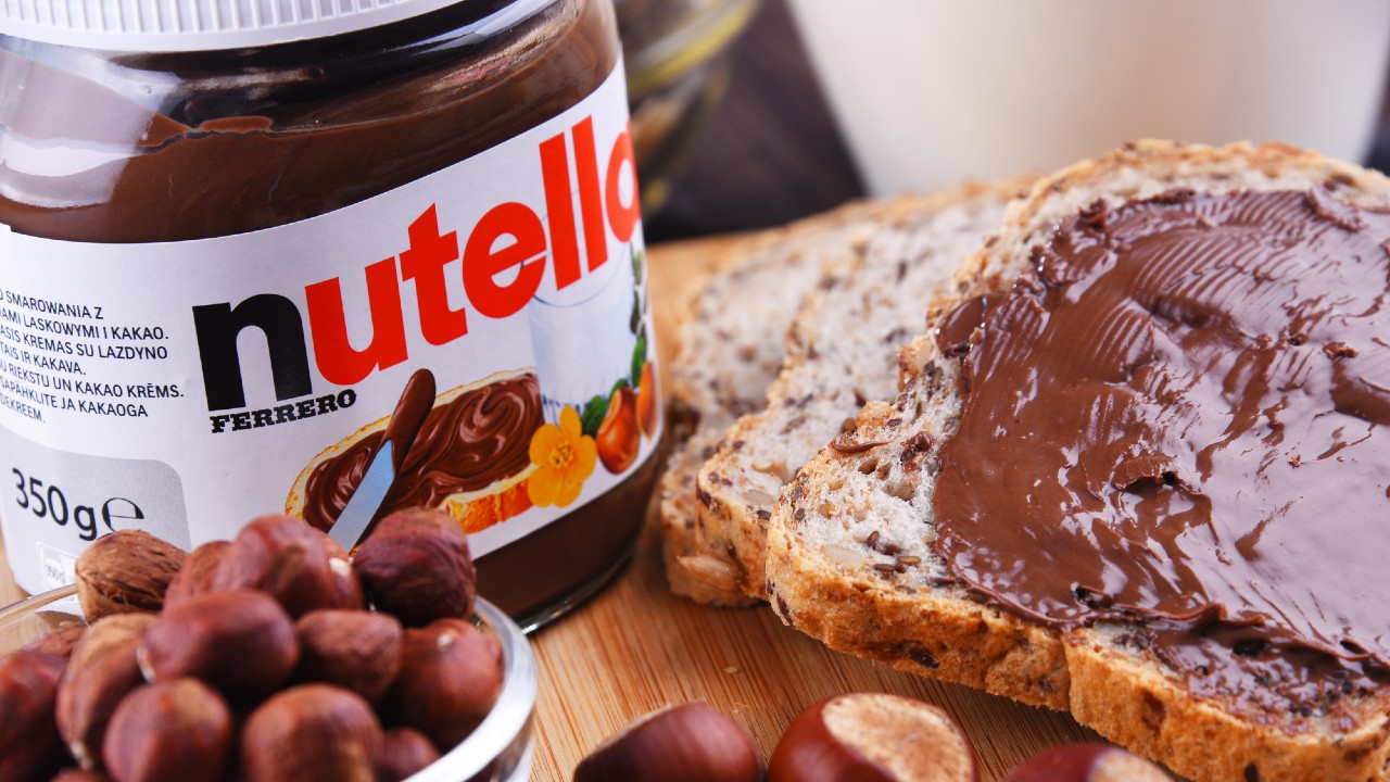 Un barattolo di Nutella con accanto due fette di pane multicereali, una delle quali spalmata di Nutella, e diverse nocciole intere (una deliziosa delizia creata dall'uomo più ricco d'Italia) sono appoggiati su una rustica superficie di legno.