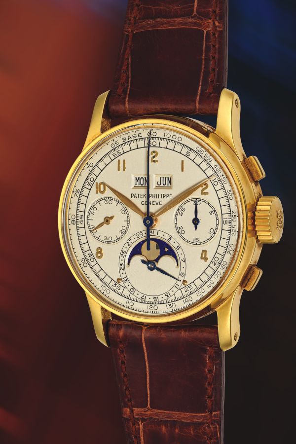 Un orologio da polso Patek Philippe in oro con cinturino in pelle marrone, esposto in un'asta Phillips, presenta un quadrante bianco con tre contatori, indicatore delle fasi lunari e finestrelle per giorno e mese. L'orologio è mostrato in primo piano su uno sfondo sfocato.