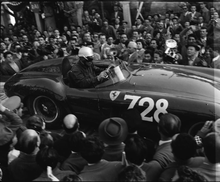 Un'auto da corsa d'epoca con il numero 728 sfreccia tra una folla festante alla Mille Miglia 2025. Il pilota, che indossa casco e occhiali protettivi, è circondato da spettatori stipati attorno al veicolo.