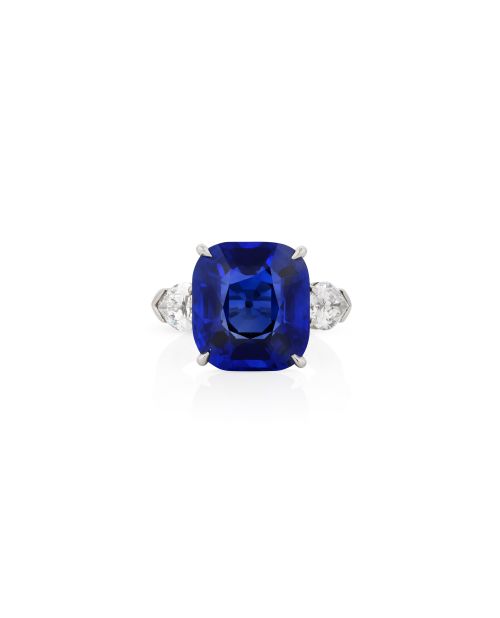 Un anello con al centro un grande zaffiro blu taglio cuscino, affiancato da due diamanti bianchi più piccoli a forma di pera su ciascun lato, incastonati in una fascia di metallo bianco che ricorda l'eleganza dell'Orologio Bulgari Serpente.