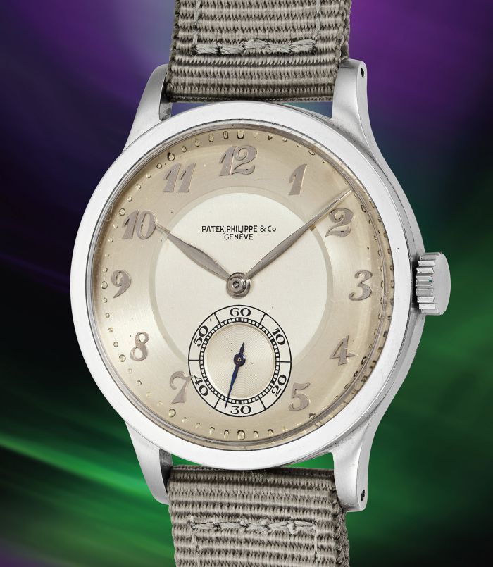 Un orologio da polso Patek Philippe in argento con quadrante beige, numeri arabi e piccolo quadrante dei secondi a ore 6 è presentato con un cinturino in tessuto leggero su uno sfondo sfocato viola e verde, che richiama l'eleganza delle aste Phillips.