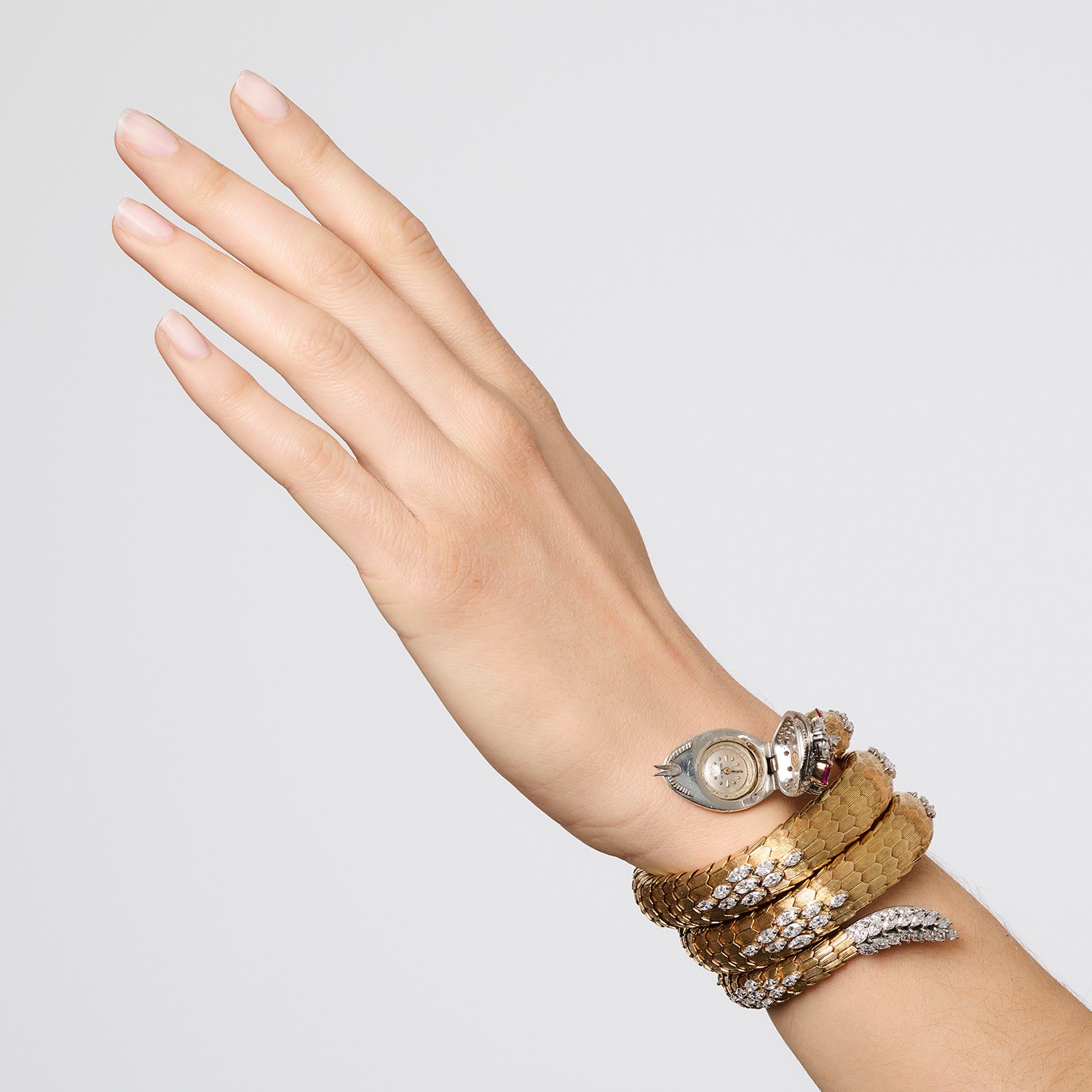 Una mano con le unghie ben curate è sollevata, adornata da un elaborato orologio Bulgari Serpente in oro e argento avvolto attorno al polso, su uno sfondo semplice e chiaro.