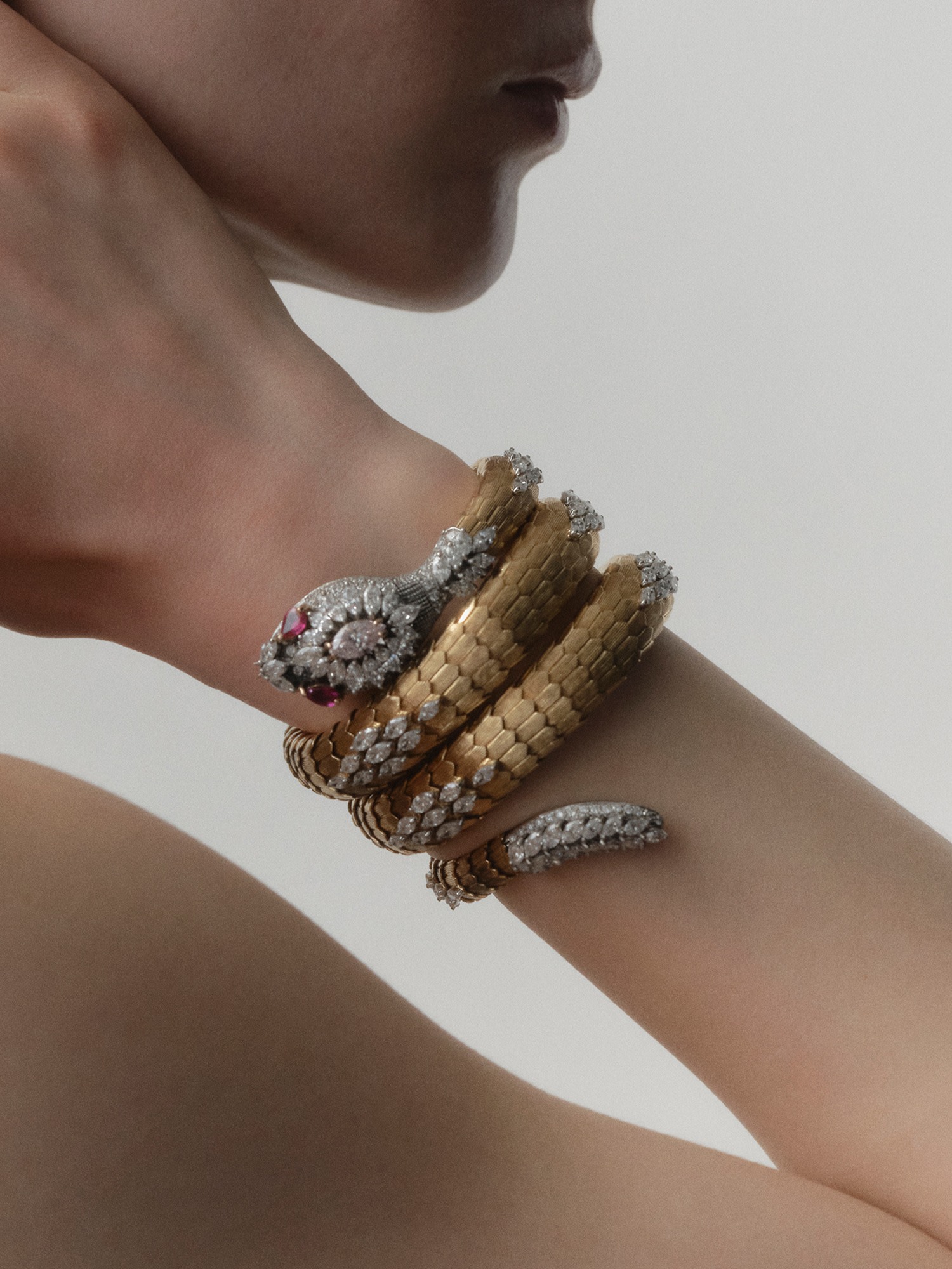 Il braccio di una persona è adornato da un bracciale a forma di serpente in oro e diamanti, che ricorda l'iconico orologio Bulgari Serpente, caratterizzato da una testa di serpente dettagliata con occhi di rubino, elegantemente avvolta attorno al polso. Lo sfondo è semplice e neutro.