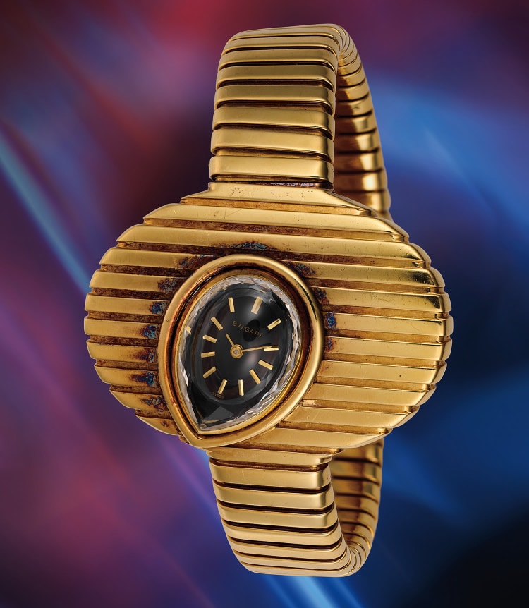 Un orologio da polso vintage Bulgari in oro con un design arrotondato e scanalato e un quadrante nero a forma di lacrima, che ricorda gli orologi visti nelle aste Phillips insieme a Patek Philippe, su uno sfondo sfumato rosso e blu.