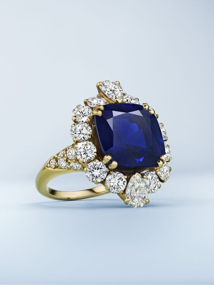 Un anello in oro con al centro un grande zaffiro blu rettangolare, circondato da un alone di diamanti bianchi rotondi e a forma di pera, incastonato su uno sfondo chiaro.