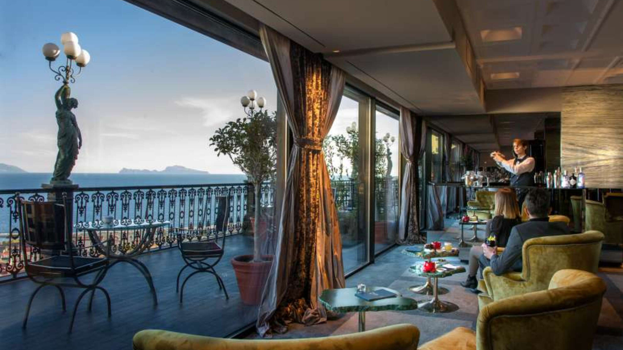 Un lussuoso salotto con poltrone gialle e tavolini si affaccia su un balcone con una splendida vista sul mare di Napoli. Le grandi finestre lasciano entrare la luce naturale, mentre il barista prepara i drink e gli avventori si rilassano e si godono il panorama.