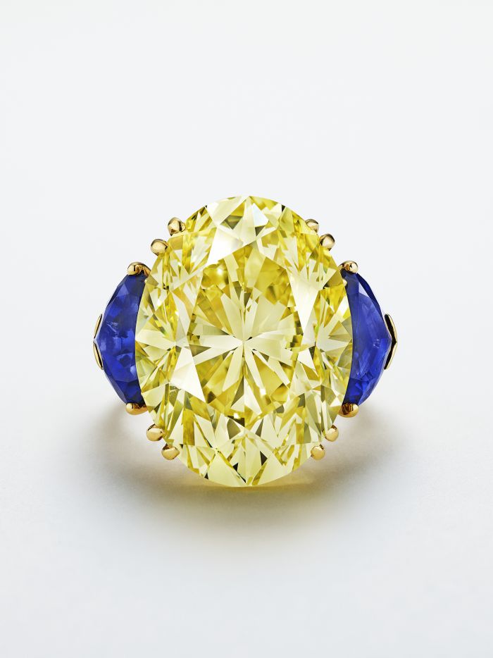 Un grande anello con diamante giallo taglio ovale, con due pietre preziose blu laterali incastonate in una fascia d'oro, su uno sfondo bianco.