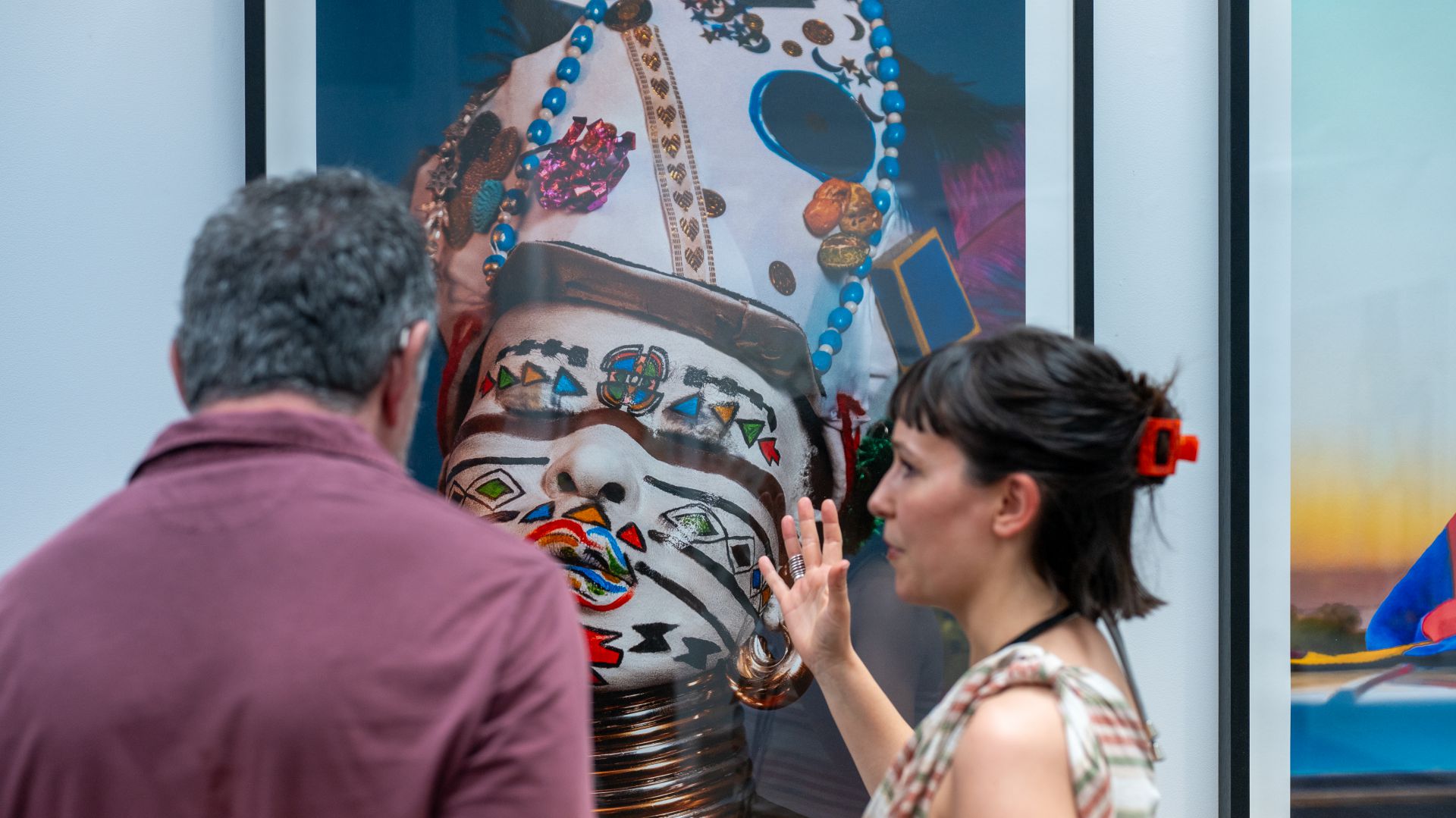 Una donna gesticola mentre parla con un uomo davanti al ritratto colorato e decorato di una persona con il volto dipinto e il copricapo esposto in una galleria d'arte.