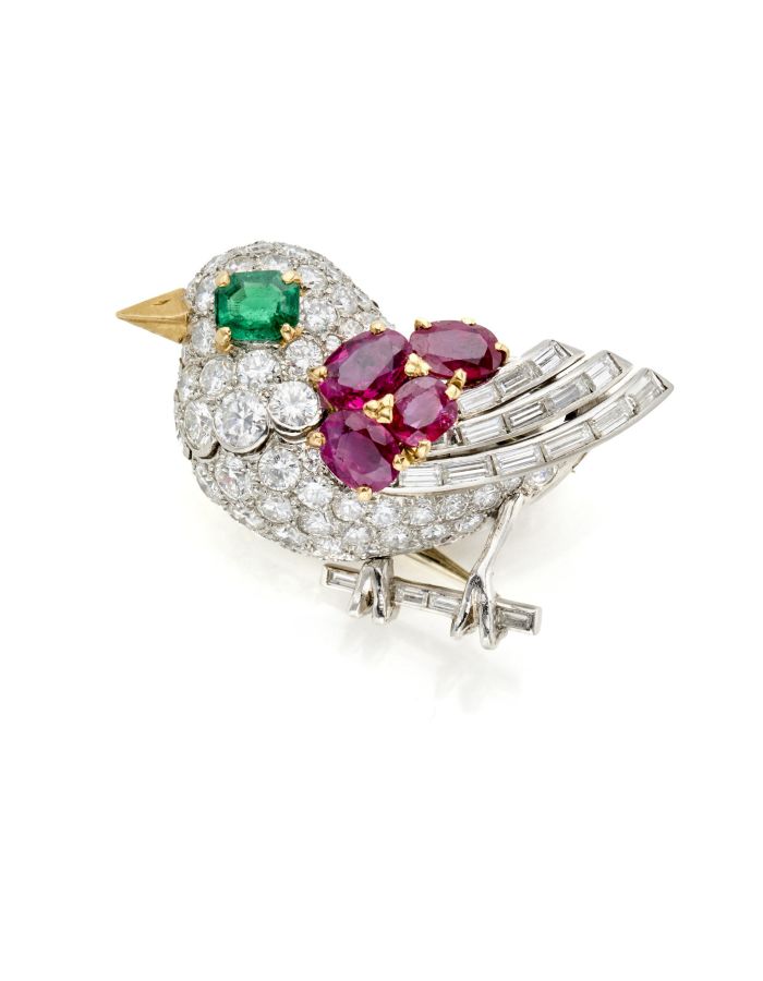 Spilla gioiello a forma di uccello con corpo tempestato di diamanti, occhio con pietra preziosa verde, becco giallo, ali di rubino e piume della coda con diamanti taglio baguette, appollaiato su un ramo d'argento.