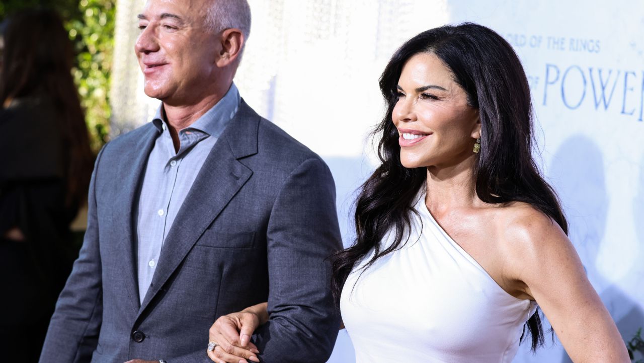 Un uomo in abito grigio e una donna in abito bianco monospalla sorridono mentre posano insieme sul tappeto rosso, con le voci del divorzio Bezos che si rincorrono mentre si trovano davanti a uno striscione promozionale chiaro.