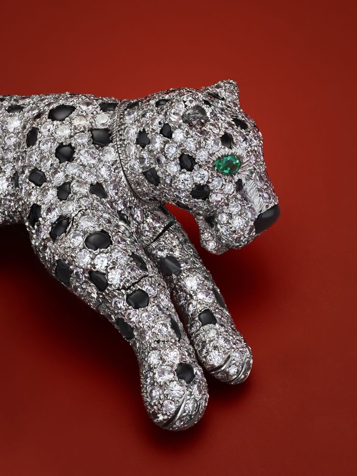 Una spilla gioiello a forma di pantera, tempestata di diamanti e pietre preziose nere, con un occhio in pietra preziosa verde, è esposta su uno sfondo rosso.