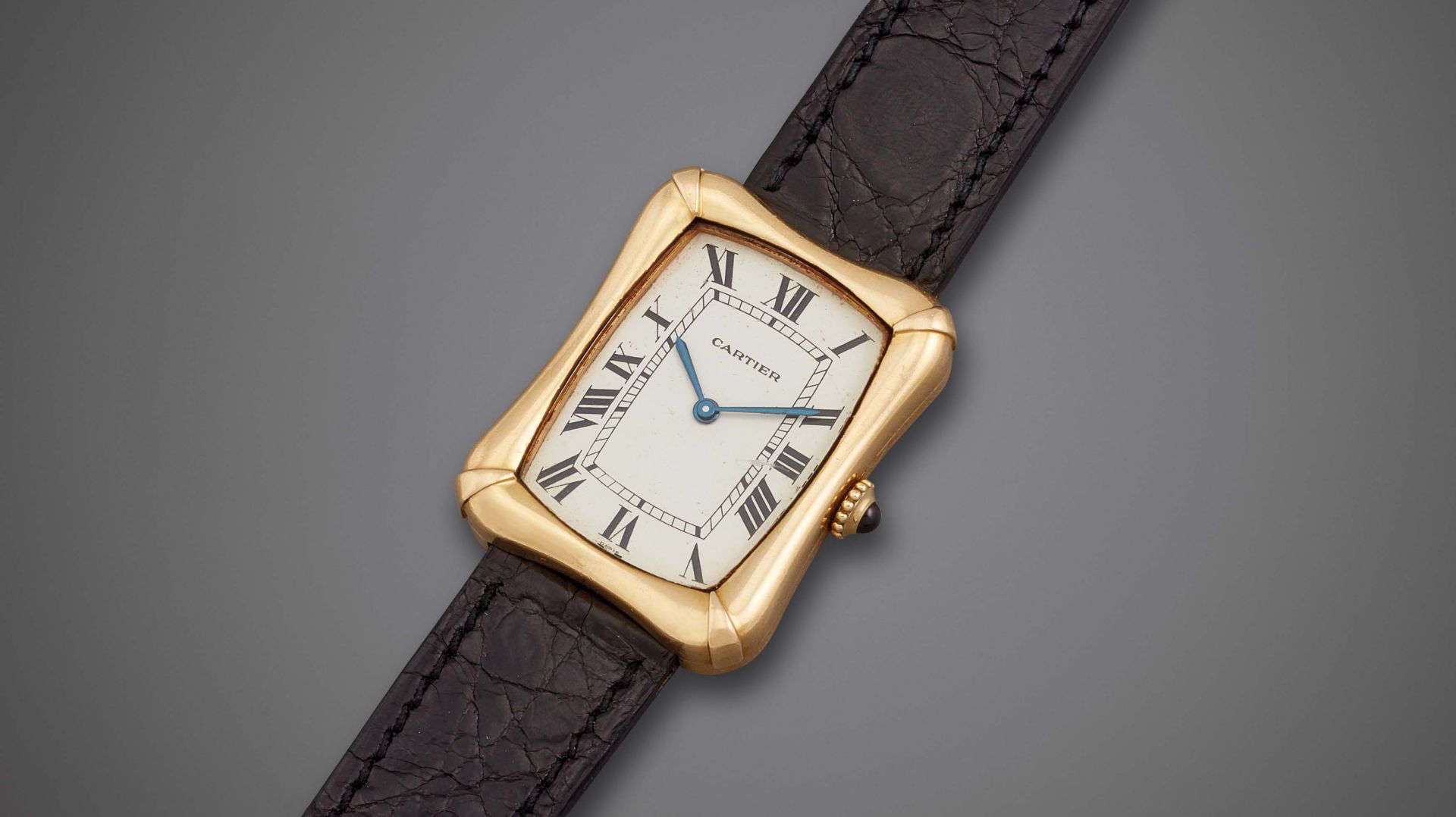 Un orologio da polso Cartier in oro con cassa rettangolare, quadrante bianco, numeri romani neri, lancette blu e cinturino in pelle nera è visualizzato su uno sfondo grigio.