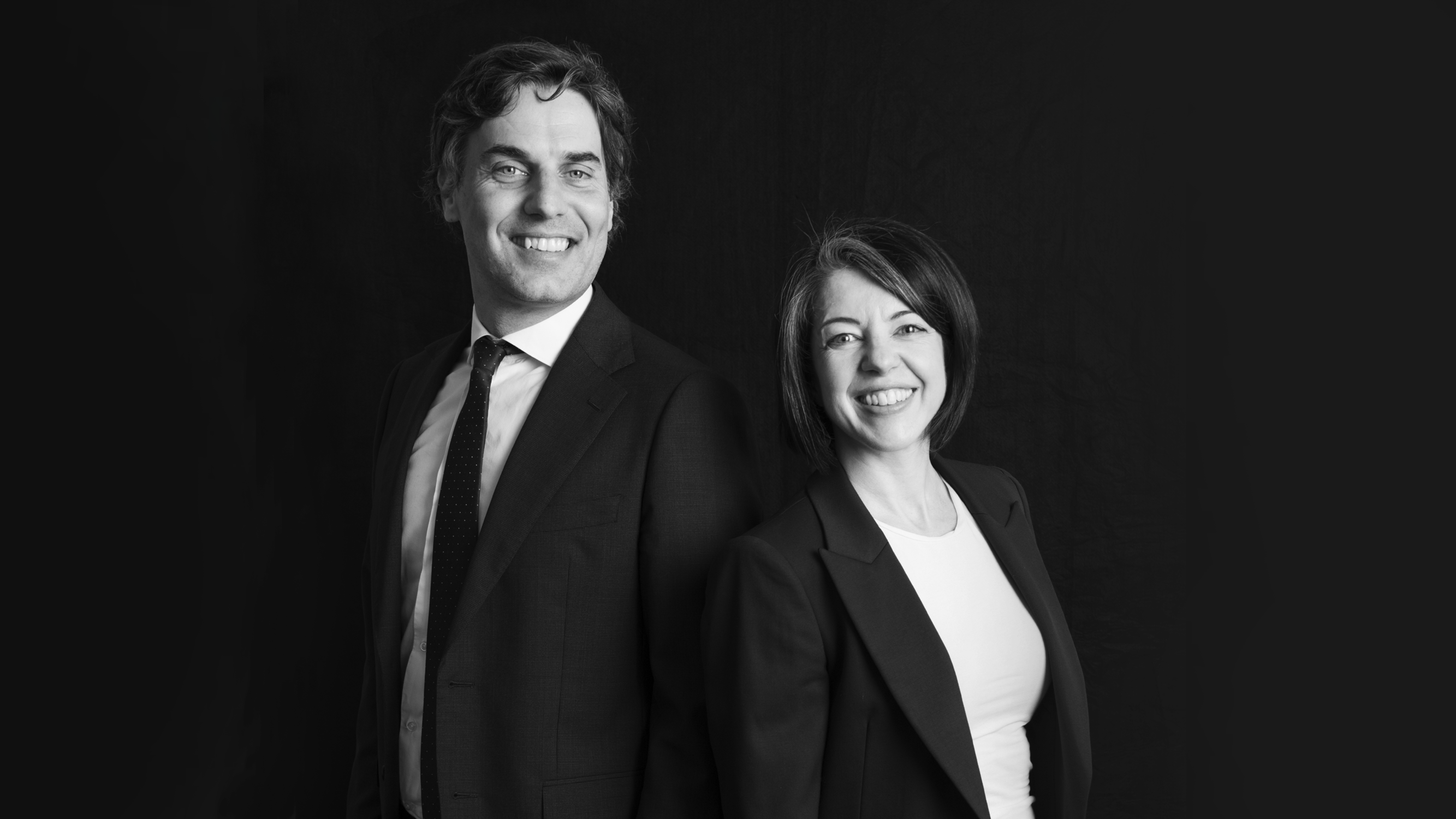Un uomo e una donna in abiti da lavoro si trovano fianco a fianco, sorridenti, su un semplice sfondo nero. L'immagine in bianco e nero evoca professionalità, perfetta per un profilo Stepstone.