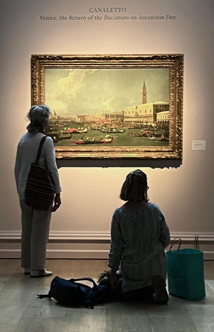 Due donne guardano un grande dipinto incorniciato di Venezia del Canaletto in un museo; una sta in piedi mentre l'altra è inginocchiata, entrambe illuminate dalla luce della galleria. Il titolo del dipinto e i recenti risultati dell'asta di Canaletto sono esposti in alto sulla parete.