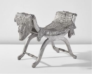 Una sedia scultorea in argento con texture a coccodrillo, caratterizzata da due teste e code di coccodrillo alle estremità, mette in mostra donne dal design unico. Le sue gambe a forma di X spiccano su un semplice sfondo bianco.