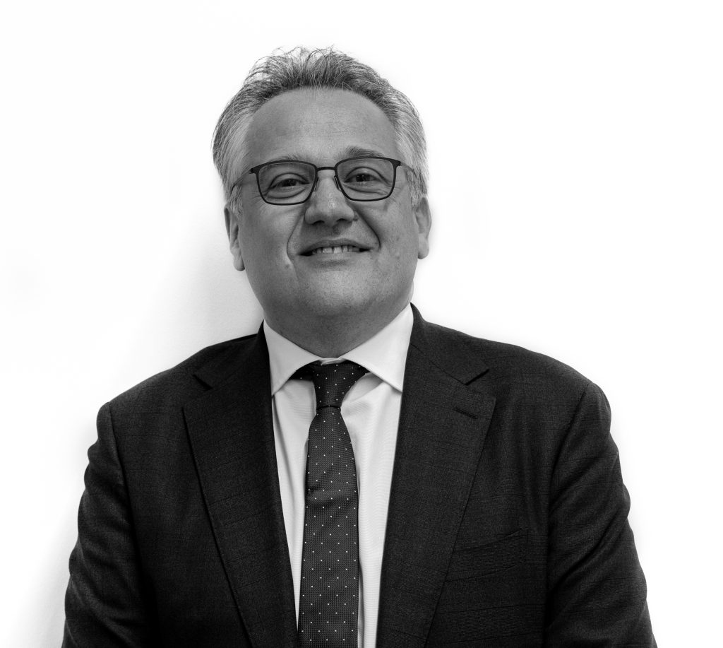 Un uomo di mezza età con i capelli corti e grigi e gli occhiali sorride. Indossa un abito scuro, una camicia bianca e una cravatta a pois, in posa su uno sfondo bianco, pronto a partecipare a eventi esclusivi di aste orologi.
