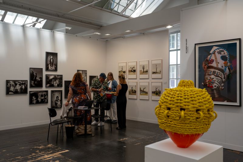 Quattro persone parlano in una galleria d'arte di Africa Basel, circondate da varie fotografie su pareti bianche. Una scultura intrecciata di colore giallo brillante si trova su un piedistallo, mentre un ritratto colorato a tecnica mista spicca sulla destra. La luce naturale riempie lo spazio.