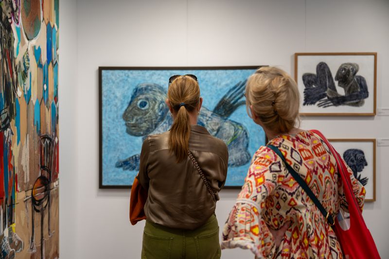 Due donne si trovano in una galleria d'arte all'Africa Basel e osservano dipinti astratti colorati, tra cui una grande opera blu con sembianze animali. Una ha la coda di cavallo; l'altra indossa un abito a fantasia e porta una borsa rossa.