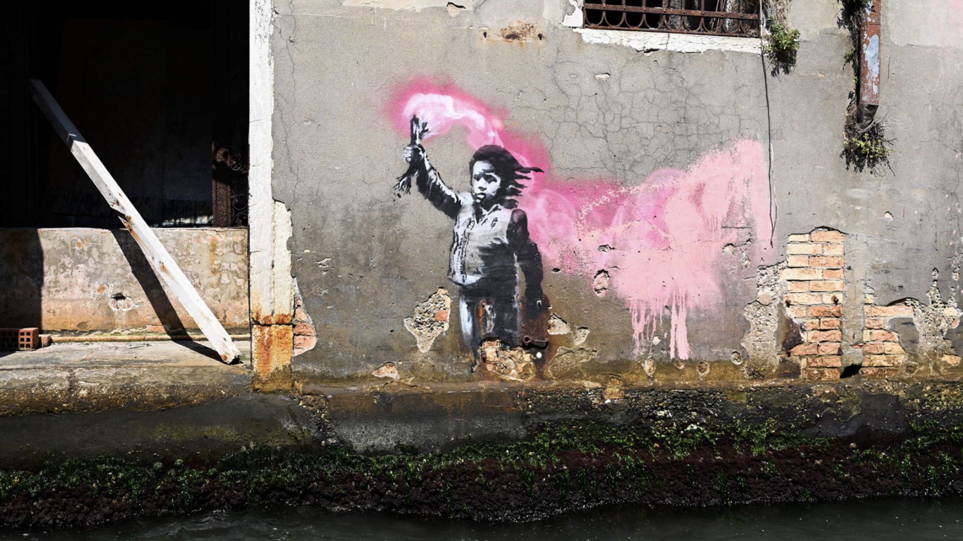 L'arte di strada su un muro danneggiato dalle intemperie raffigura un bambino con un giubbotto di salvataggio che tiene in mano un razzo di segnalazione rosa, con la vernice rosa che si allunga sulla destra. Il muro è crepato, con muschio in basso e finestre sbarrate in alto.