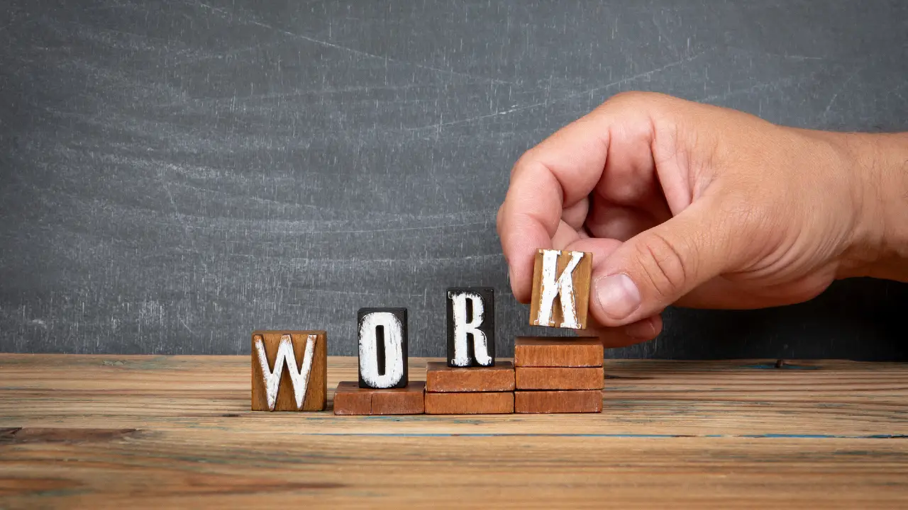 Una mano posiziona un blocco di legno con la lettera "K" accanto a blocchi con la scritta "WORK", disposti in modo graduale su una superficie di legno con sfondo a lavagna.