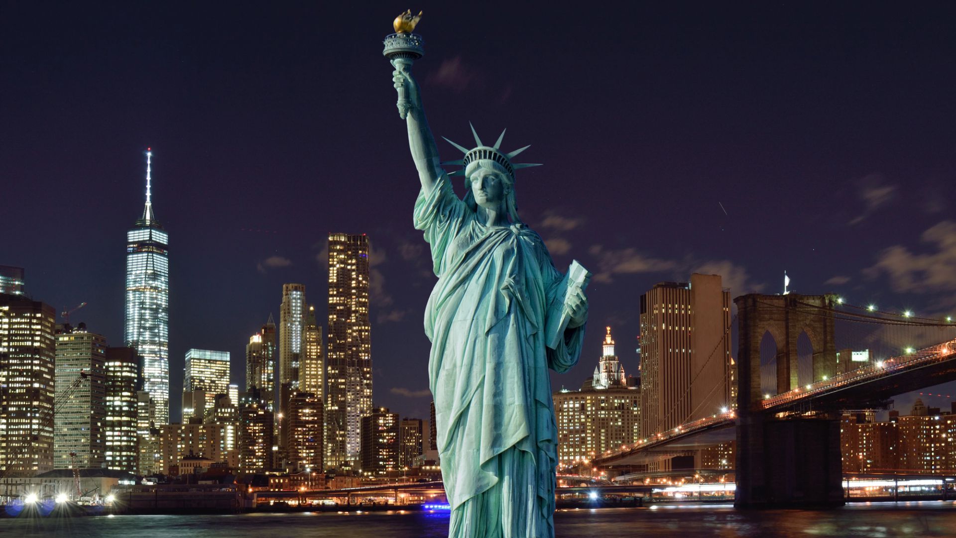 La Statua della Libertà illuminata di notte, di fronte allo skyline di New York, con i grattacieli e il ponte di Brooklyn visibili sotto il cielo scuro.