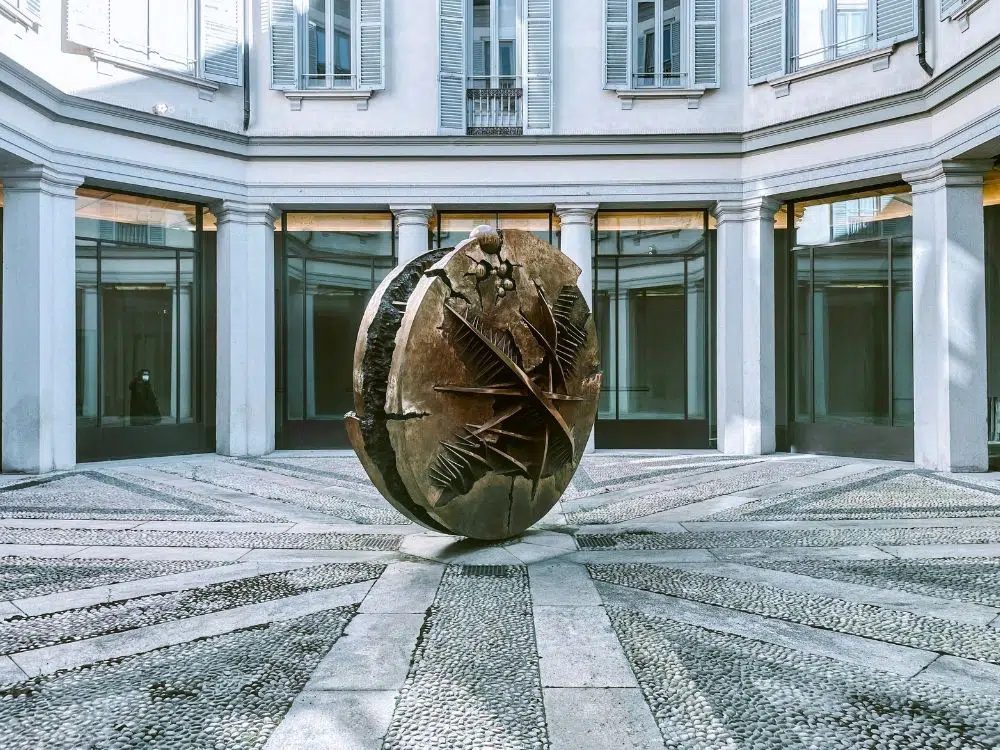 Una grande scultura a sfera in bronzo di Arnaldo Pomodoro, ornata da motivi geometrici e astratti, si trova al centro di un cortile circolare in pietra, circondato da un edificio bianco con alte finestre e colonne.