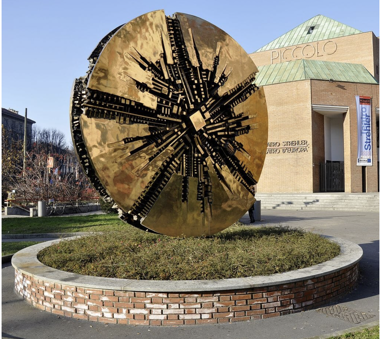 Una grande scultura circolare in bronzo di Arnaldo Pomodoro, caratterizzata da intricati ritagli geometrici, si trova su una collinetta erbosa circondata da un bordo in mattoni di fronte all'edificio moderno denominato "Piccolo Teatro Strehler".