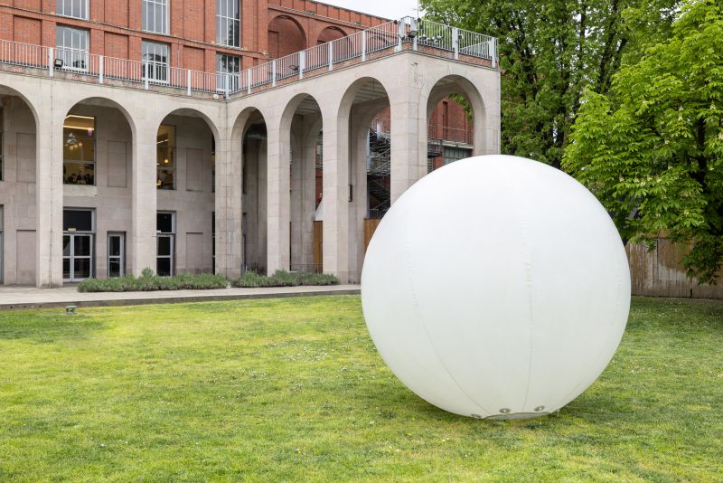 Una grande sfera bianca si trova su un prato erboso di fronte a un edificio in pietra con archi alti e grandi finestre, che evoca il fascino contemporaneo della Triennale di Gucci. Sulla destra sono visibili alberi con foglie verdi.