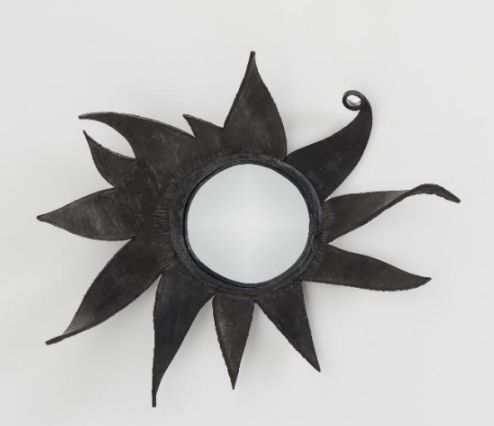 Uno specchio rotondo con una cornice decorativa in metallo nero a forma di sole astratto, con raggi irregolari e appuntiti e una punta arricciata su un raggio - un design unico Donne Asta - incastonato su uno sfondo bianco.
