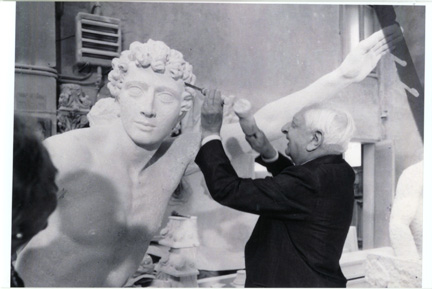 Un uomo anziano in giacca e cravatta, che fa parte della Triennale di Gucci, restaura con cura una grande statua classica, utilizzando un piccolo strumento per lavorare sul volto della statua in uno studio al chiuso.
