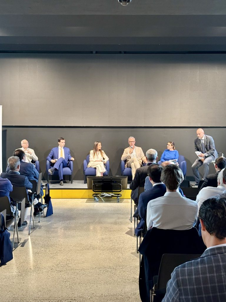 Cinque relatori sono seduti su un palco e discutono davanti a un pubblico in una moderna sala conferenze. Il pubblico è seduto e rivolto con attenzione verso i relatori. Lo sfondo è semplice e scuro.