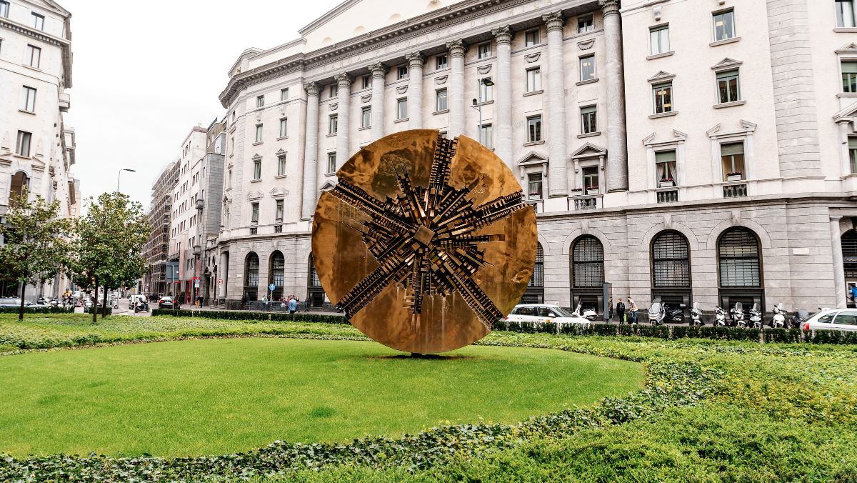 Una grande scultura circolare in bronzo con intricati motivi geometrici si trova in un'area erbosa circondata da siepi, di fronte a un grande edificio storico con finestre ad arco e decorazioni in pietra.