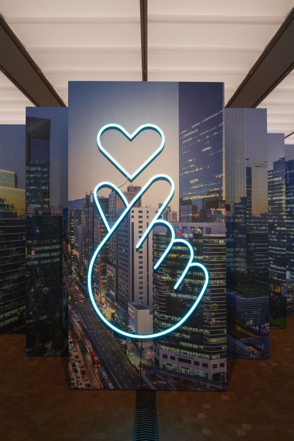 Un'insegna al neon raffigurante una mano che fa il gesto del cuore con un cuore sopra di essa, sullo sfondo di un paesaggio urbano moderno, che cattura l'energia vibrante e l'influenza hallyu nelle strade urbane di oggi.