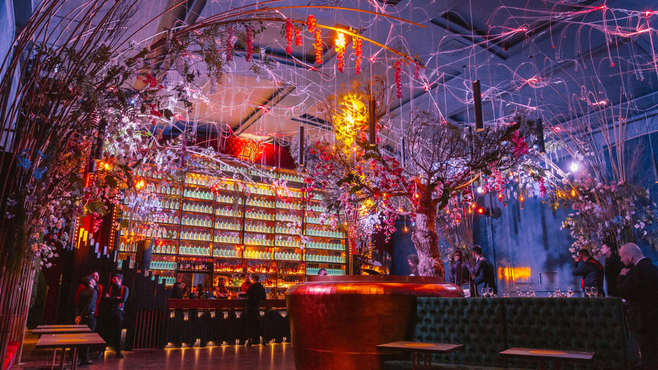 Un vivace bar di Napoli con luci colorate, scaffali di bottiglie e scenografiche decorazioni floreali, tra cui un grande albero centrale. Le persone sono riunite intorno, creando una scena vivace e suggestiva.