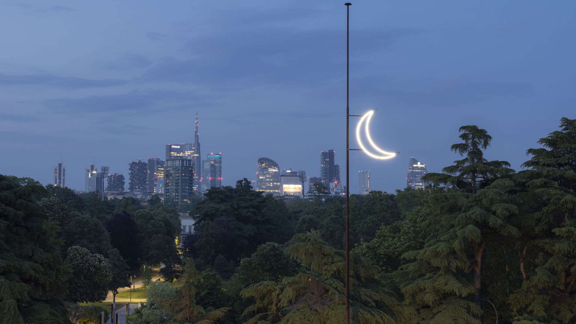 Lo skyline di una città al tramonto è caratterizzato da edifici moderni e da un parco lussureggiante, con una lampada a forma di mezzaluna che brilla come una luna: una scena che ricorda l'eleganza delle installazioni della Triennale di Gucci.