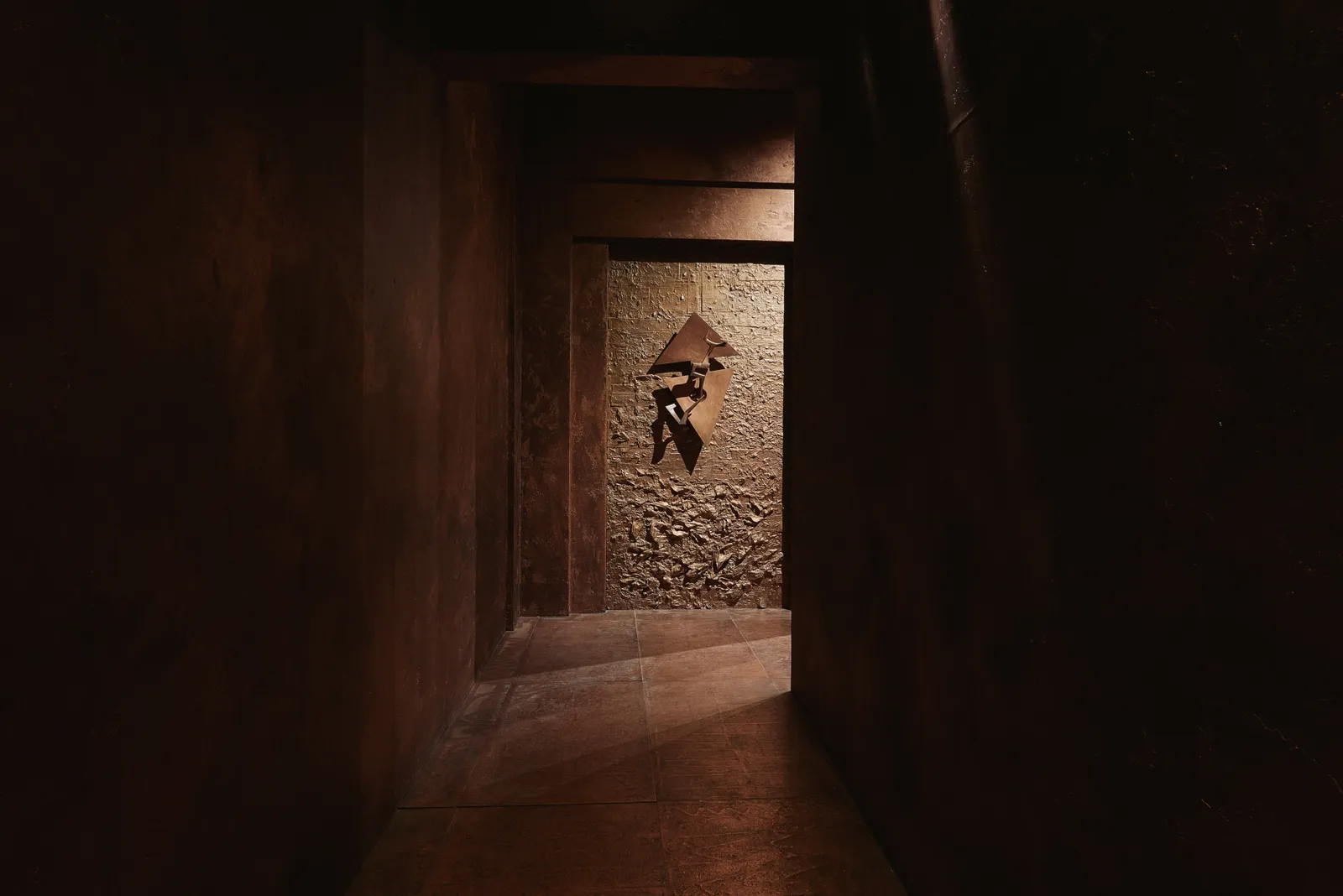 Un corridoio stretto e poco illuminato, con pareti e pavimento di colore marrone, conduce a una parete con una scultura geometrica astratta in metallo che ricorda Arnaldo Pomodoro. La luce calda evidenzia la superficie ruvida e l'opera d'arte in fondo.