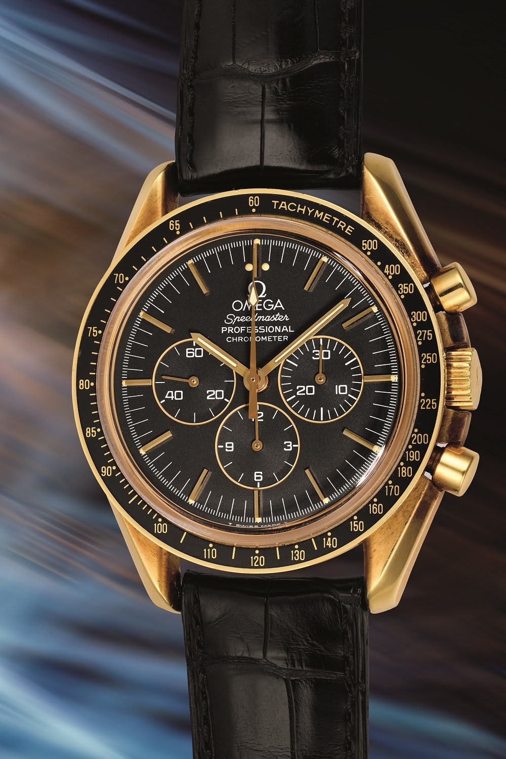 Un orologio da polso Omega Speedmaster Professional in oro con quadrante nero, tre quadranti secondari e cinturino in pelle nera, esposto su uno sfondo sfocato e dinamico: un'elegante alternativa a Rolex o Patek alla tua prossima asta.