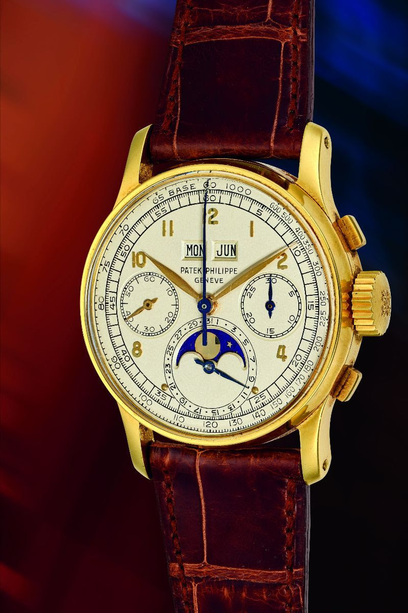 Un orologio da polso Patek Philippe in oro con cinturino in pelle marrone, dotato di tre quadranti secondari, indicatore delle fasi lunari e finestrelle per giorno e mese su un quadrante bianco: un segnatempo raffinato, perfetto per qualsiasi asta, su uno sfondo sfumato rosso e blu.