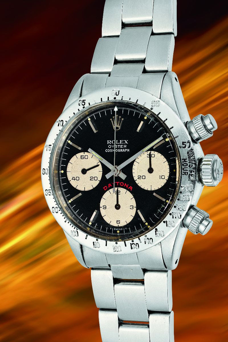 Un orologio Rolex Cosmograph Daytona d'epoca con quadrante nero, tre quadranti secondari e bracciale in acciaio inossidabile è esposto su uno sfondo sfocato dinamico arancione e giallo, che mette in risalto l'eleganza classica del Rolex Patek Asta.